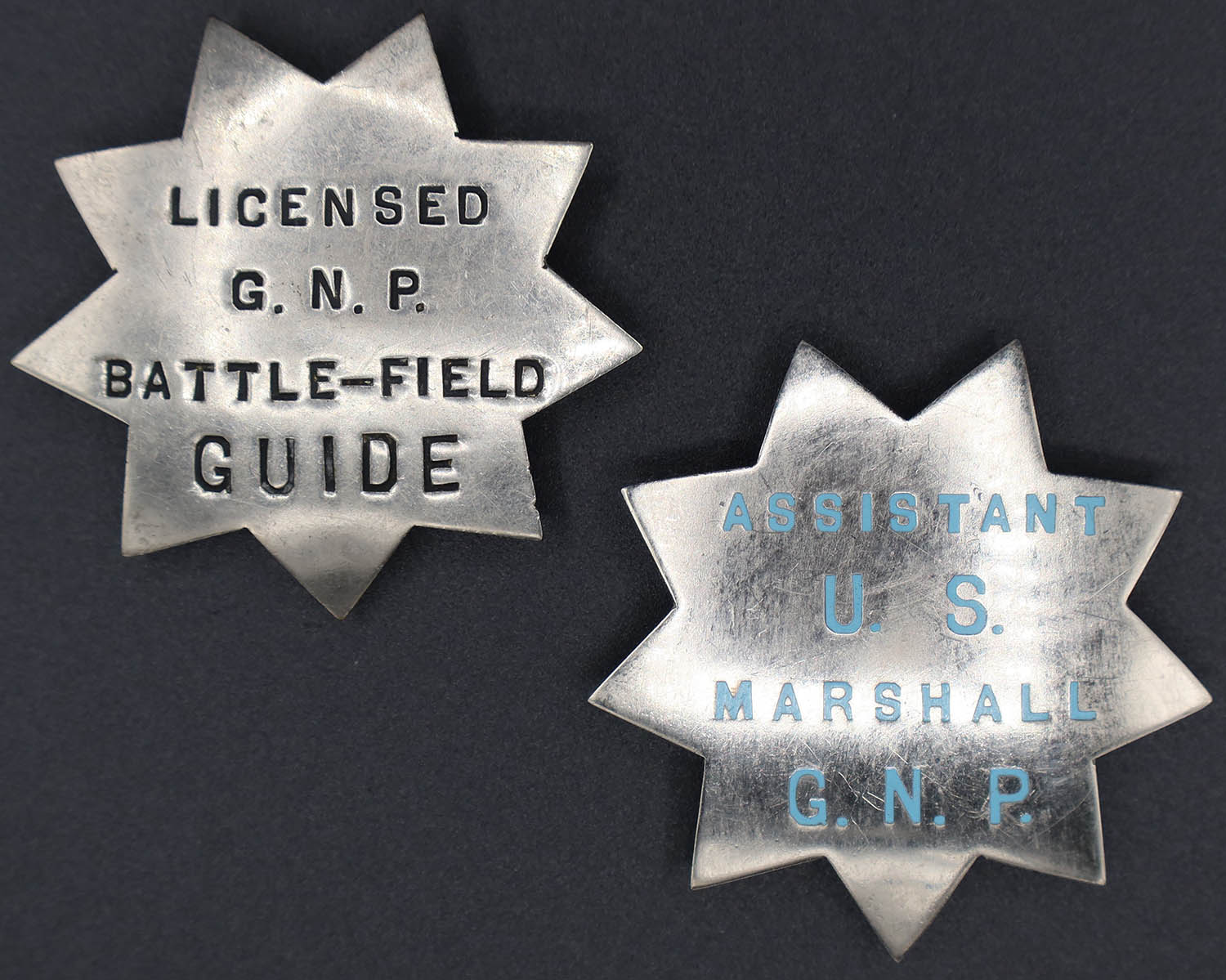 Two silver badges shaped as 9-point stars. One is marked with black letters for a licensed guide. The other in blue for an assistant marshal.